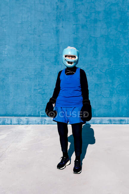Mujer madura de cuerpo entero en ropa deportiva y guantes de boxeo de pie con casco contra la pared azul y mirando a la cámara - foto de stock