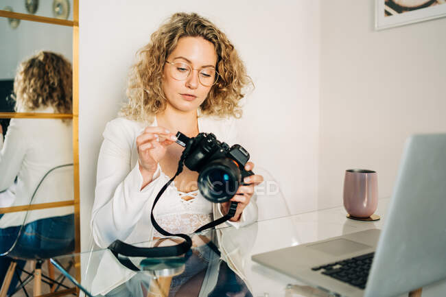 Серйозна молода жінка з кучерявим світлим волоссям у стильному вбранні та окулярах вставляє SD-карту в фотоапарат після перенесення файлів на ноутбук вдома — стокове фото