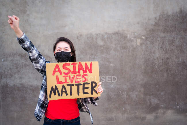 Mujer étnica en máscara y con cartel de cartón con inscripción Asian Lives Matter protestando con el brazo levantado en la calle de la ciudad y mirando hacia otro lado - foto de stock