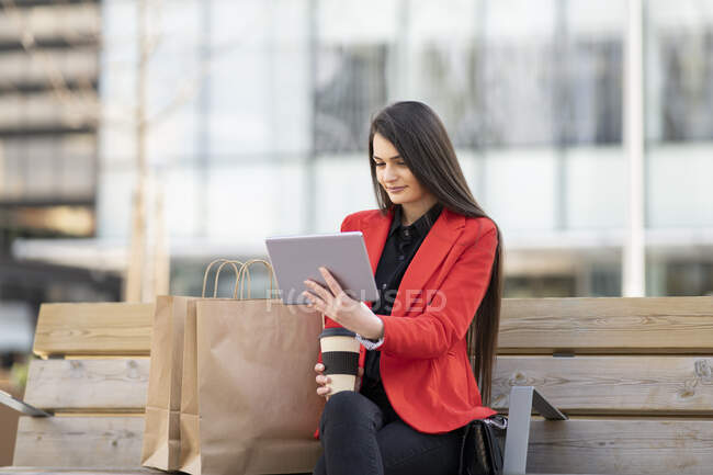 Entzückte Käuferin sitzt mit Papiereinkaufstüten auf Bank und schaut sich Video auf Tablet in der Stadt an — Stockfoto