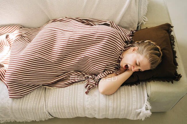 From above of young sleepy female yawning while lying under plaid on soft sofa at home — Stock Photo