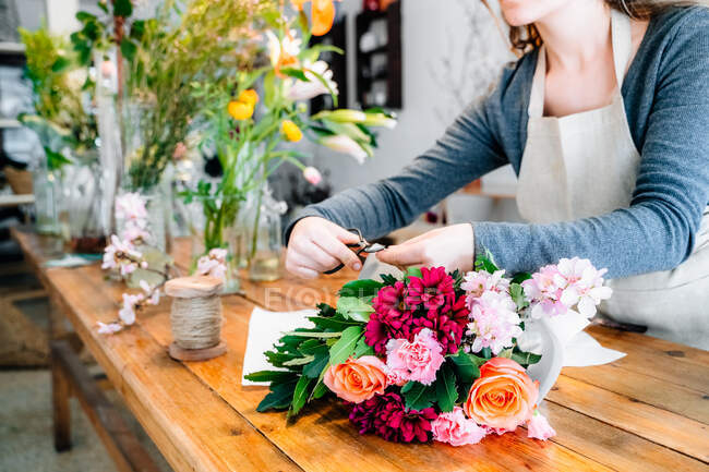 Crop unrecognizable young female florist with long wavy hair in casual clothes and apron arranging elegant bouquet of roses and assorted flowers in store — Stock Photo