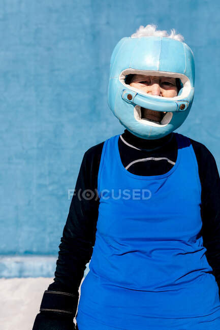 Femme mûre en vêtements de sport et gants de boxe debout avec casque contre le mur bleu et regardant la caméra — Photo de stock