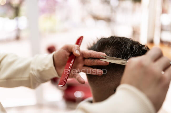 Crop anonymous male stylist with trimmer cutting hair of client in cape in barbershop — Photo de stock