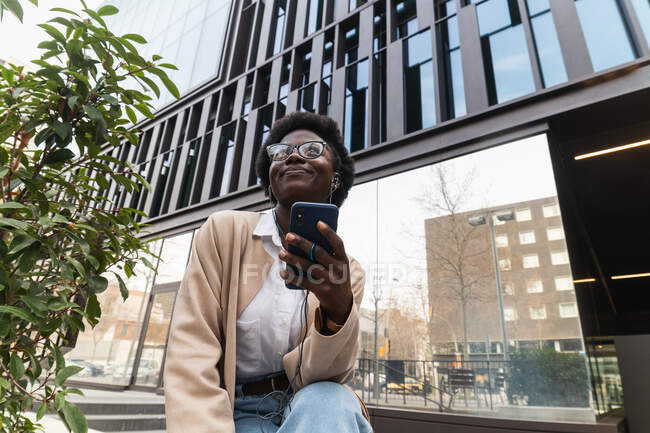 From below of smiling African American female entrepreneur sitting near office building in city and surfing Internet on smartphone — Stock Photo