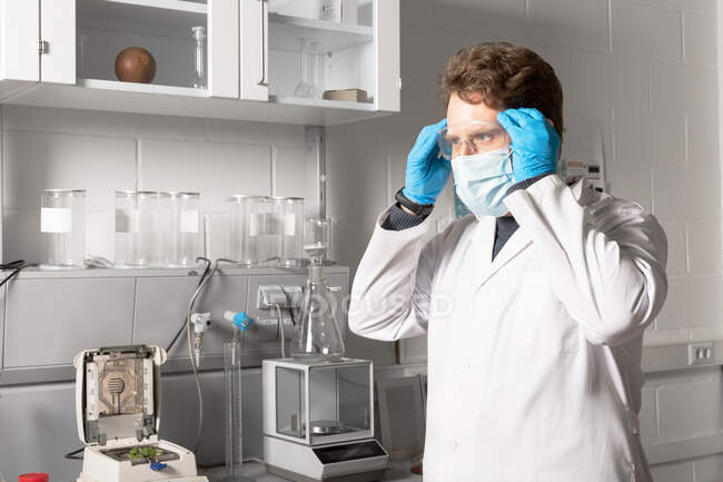Químico masculino en máscara estéril y guantes mirando hacia adelante contra el analizador de humedad y el equilibrio analítico en el laboratorio de cannabis - foto de stock