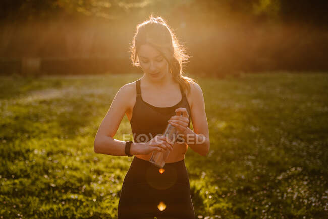 Young sportswoman with bottle of water watching pulse rate on wearable tracker during break from training in back lit — Stock Photo