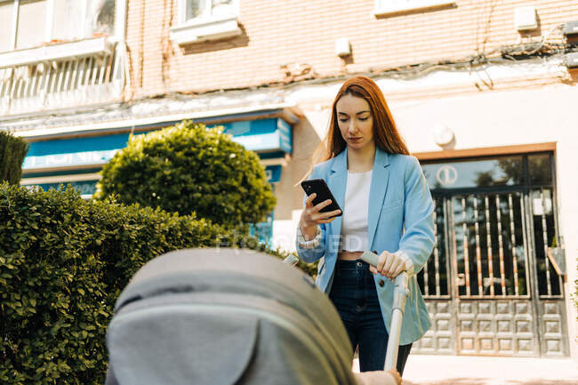 Concentrated young mother in casual wear browsing mobile phone while standing on sunny summer street with baby stroller — Stock Photo