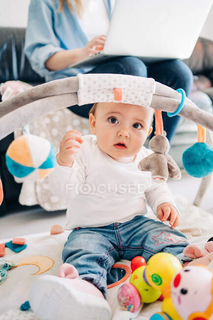 Entzückendes kleines Baby, das mit Spielzeug auf dem Boden spielt und wegschaut, während es neben der Mutter sitzt und im hellen Wohnzimmer Netbook blättert — Stockfoto