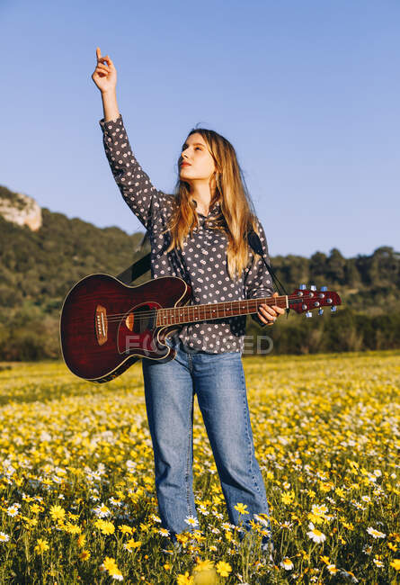 Продумана молода хіпстерка стоїть на лузі, вказуючи на небо в сільській місцевості і граючи на гітарі під час літнього сонячного світла — стокове фото