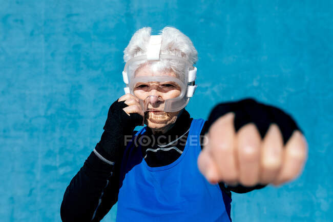 Focused mature female boxer in helmet and hand wraps punching air against blue wall in outdoor sports center — Stock Photo