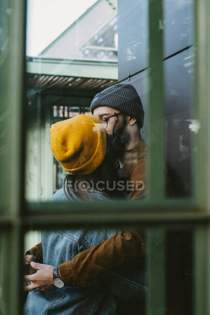 Attraverso vetro vista laterale di elegante coppia di hipster in piedi edificio pulito e baciare — Foto stock