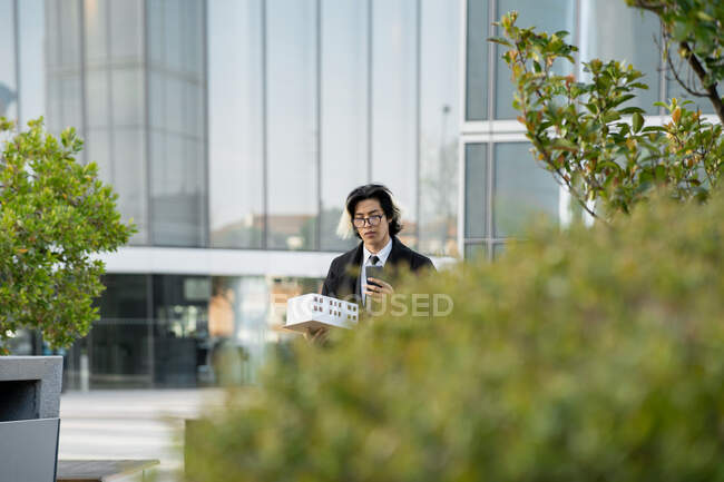 Jovem executivo étnico masculino em óculos com maquete de casa e celular olhando para a tela na cidade — Fotografia de Stock