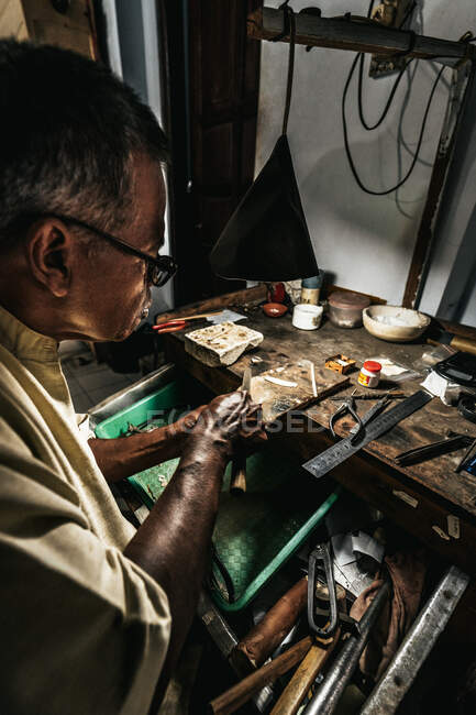 Сверху концентрированный пожилой мужчина-ювелир в повседневной одежде и очках, затачивающий инструменты, работая за деревянным столом в традиционной мастерской — стоковое фото