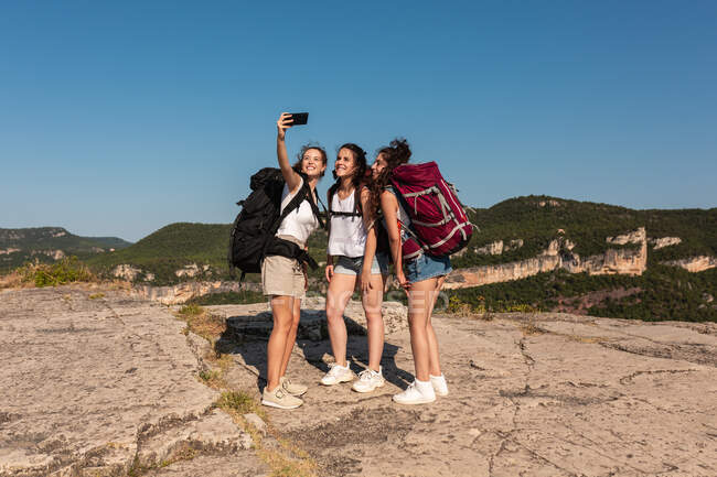 Gesellschaft fröhlicher Backpackerinnen, die auf einem Hügel im Hochland stehen und beim Wandern im Sommer Selbstaufnahmen mit dem Smartphone machen — Stockfoto