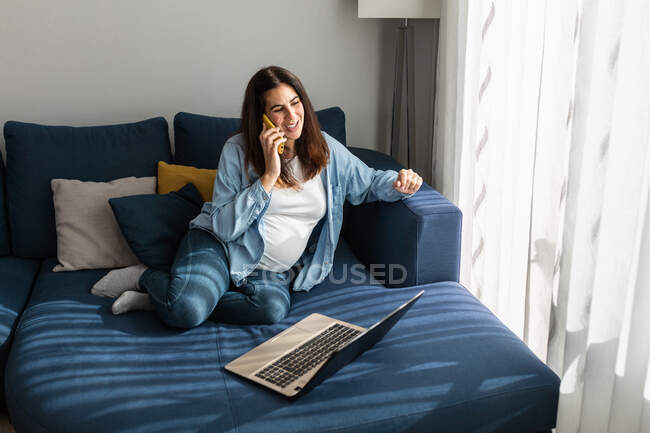 Freelancer grávida sentada no sofá com laptop na sala de estar e falando no celular enquanto discute projeto remoto e trabalha em casa — Fotografia de Stock