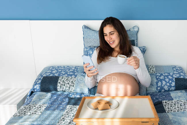 Веселая беременная женщина сидит на кровати утром и завтракает во время просмотра смартфона — стоковое фото