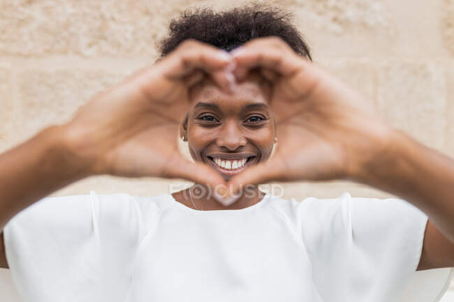 Happy young African American female in white blouse showing heart sign with hands and looking at camera with toothy smile while standing against uneven wall — Stock Photo