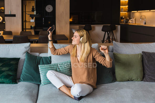 Mujer optimista sentada en el sofá y escuchando música en auriculares mientras disfruta de canciones con las manos levantadas - foto de stock