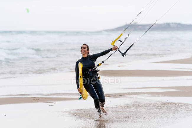 Athlète féminine en combinaison avec barre de contrôle regardant loin sur la rive sablonneuse contre l'océan mousseux après avoir pratiqué le kitesurf — Photo de stock