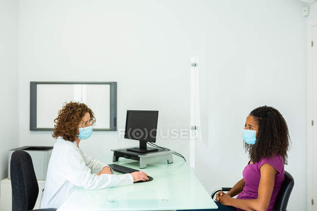 Vista laterale della donna afroamericana in maschera durante l'appuntamento seduta alla scrivania del medico donna protetta da uno schermo in vetro plexiglas presso la moderna clinica durante l'epidemia di coronavirus — Foto stock