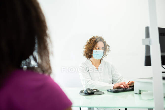 Зіпсована незнайома афро-американська жінка під час призначення сидячи в жіночому лікаря маску з офісним столом захищені екраном перспективного скла в сучасній клініці під час спалаху коронавірусу. — стокове фото