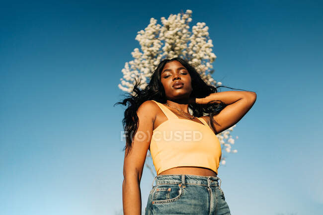 Low angle of dreamy African American female standing in blooming spring park and enjoying sunny weather with closed eyes — Stock Photo