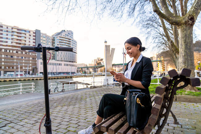 Mujer empresaria étnica sonriente sentada en un banco de madera con las piernas cruzadas hablando por teléfono celular mientras mira hacia otro lado en el banco de la ciudad contra el scooter - foto de stock