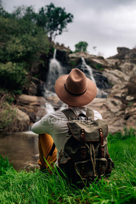 Vista posterior mochilero masculino anónimo en sombrero disfrutando de la vista de la cascada de streaming de roca rugosa en la naturaleza verde - foto de stock