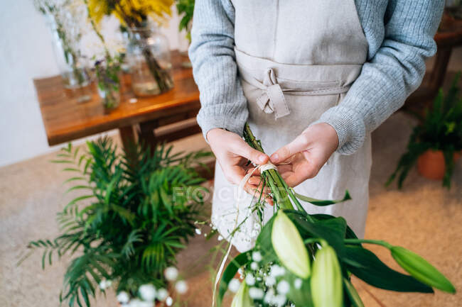 Cropped unrecognizable female florist in casual clothes and apron tying delicate bouquet of orange lily and white gypsophila in floral shop — Stock Photo