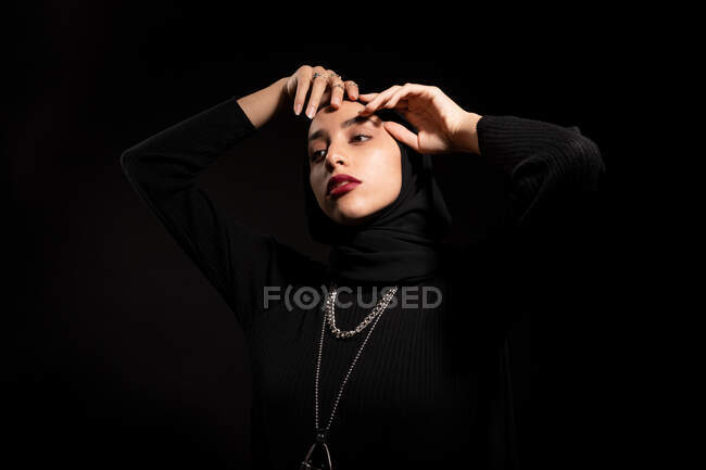 Attraente giovane donna islamica vestita di nero e hijab che tocca delicatamente il viso e distoglie lo sguardo — Foto stock