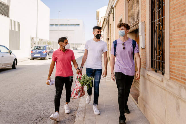 Cuerpo completo de elegantes amigos masculinos multirraciales jóvenes en ropa casual y máscaras protectoras caminando por la calle de la ciudad después de comprar en el supermercado durante la pandemia de COVID - foto de stock