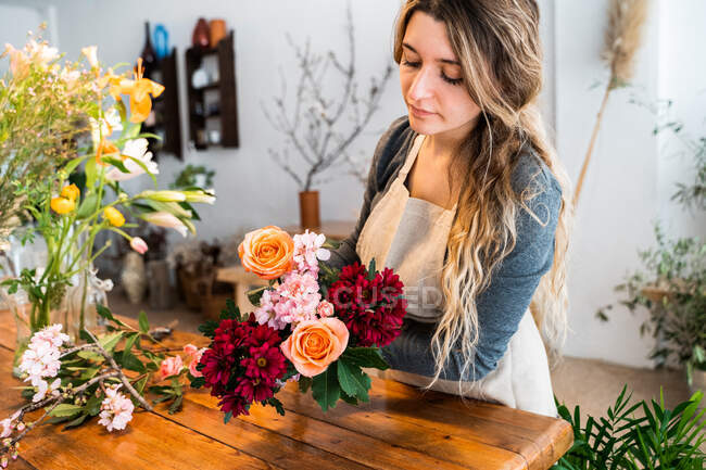 Florista femenina joven concentrada con el pelo ondulado largo en ropa casual y delantal que arregla el ramo elegante de rosas y flores surtidos en la tienda - foto de stock