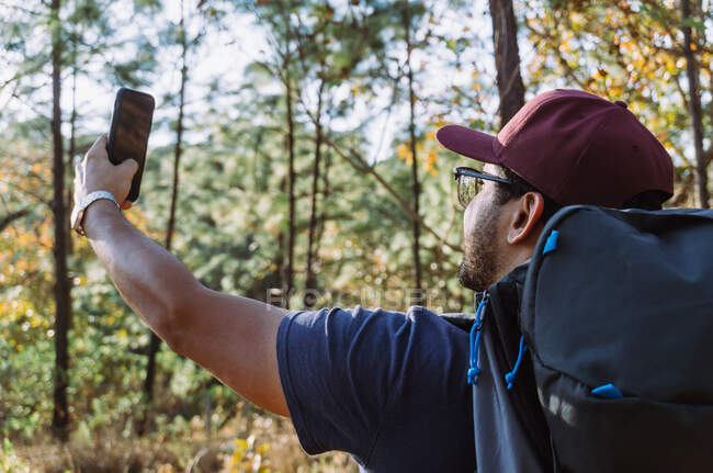 Side view of young ethnic male hiker in casual wear and cap with backpack taking a self portrait with his mobile phone in lush summer woodland — Stock Photo