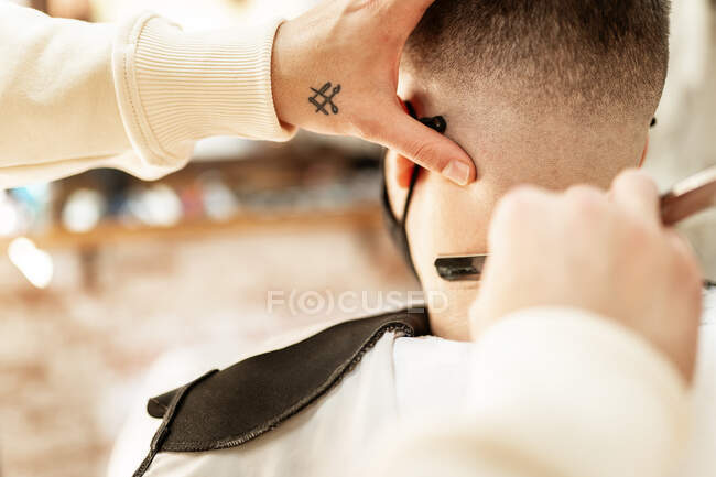 Crop anonymous hairdresser shaving man in mask with straight razor in barbershop on blurred background — Fotografia de Stock