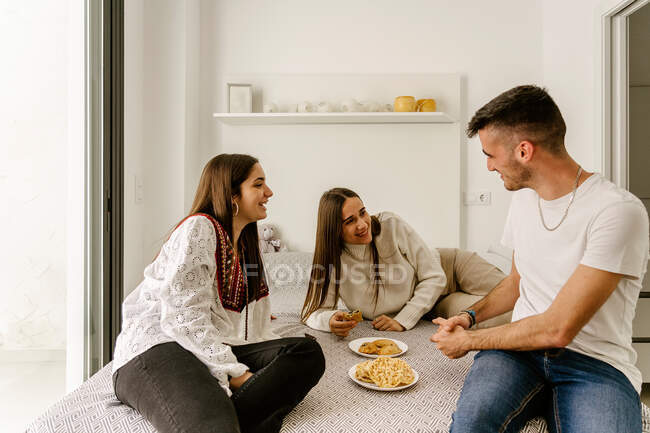 Cheerful young ethnic male and female friends in casual clothes relaxing on bed and eating yummy pastries during weekend at home — Fotografia de Stock