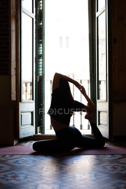Vista lateral de una delgada hembra sentada en una esterilla en Eka Pada Rajakapotasana y practicando yoga en casa - foto de stock