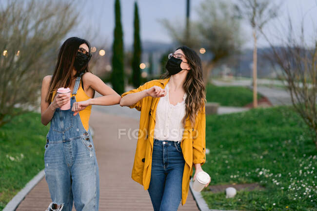 Zufriedene beste Freundinnen in Freizeitkleidung und Gesichtsmasken mit heißen Getränken im Becher berühren die Ellbogen, während sie sich in der Stadt beim Ausbruch des Coronavirus anschauen — Stockfoto