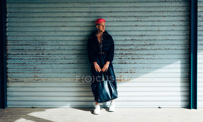 Transsexual man in trendy wear with handbag and pink hair looking away against weathered wall on sunny day — Stock Photo