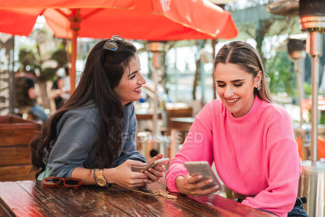 Оптимістичні молоді друзі-жінки в повсякденному вбранні використовують смартфони і обговорюють новини, сидячи за столом у відкритому ресторані Веранди в сонячний день — стокове фото