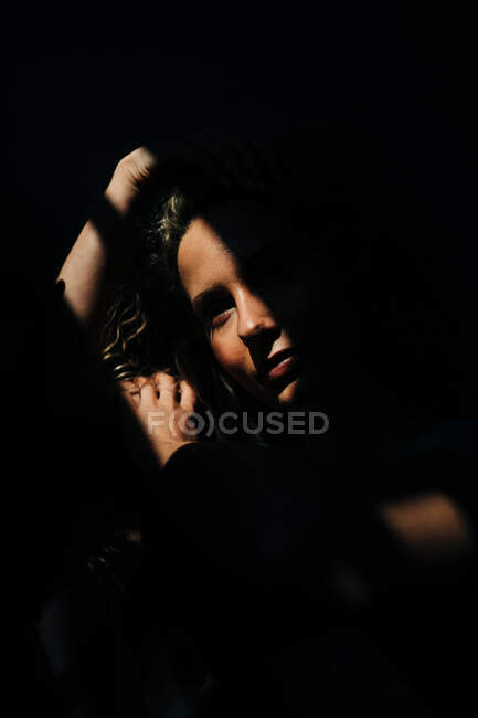 Calm young female touching hair and looking away while standing in dark room in shadows — Stock Photo