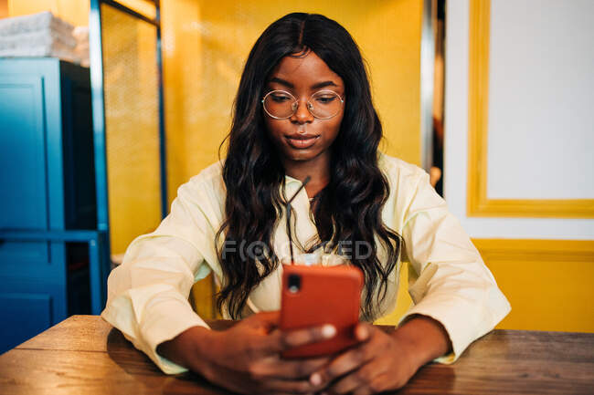 Encantadora mujer afroamericana sentada en la mesa navegando por el teléfono móvil mientras mira la pantalla - foto de stock