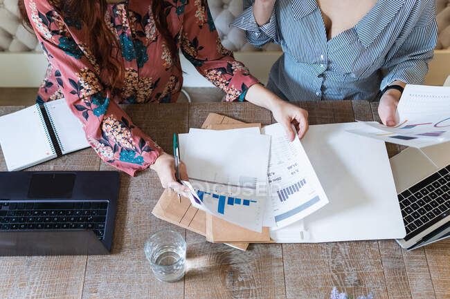 Desde arriba, las compañeras de trabajo se reúnen en el escritorio con computadoras portátiles y analizan diagramas mientras trabajan juntas en un espacio de trabajo moderno - foto de stock