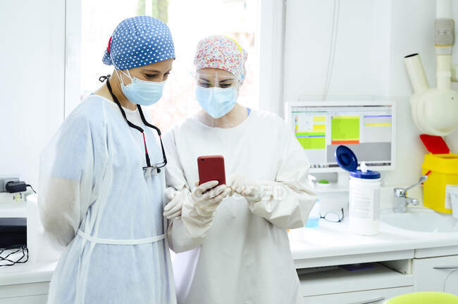 Female surgeons in medical cloth caps surfing internet on cellphone against desktop computer in light hospital — Stock Photo