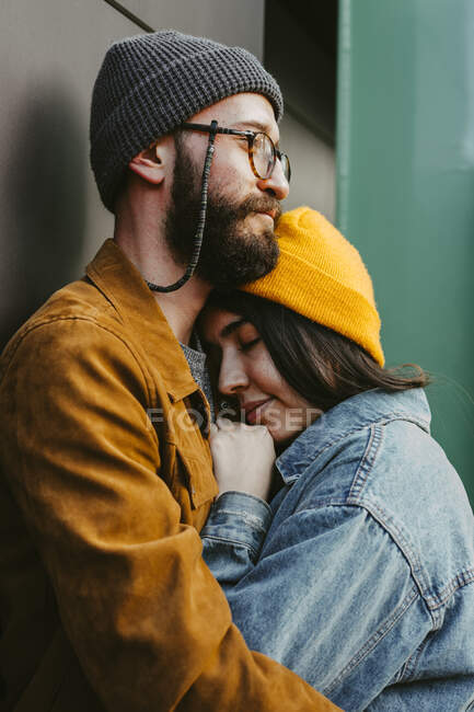 Вид збоку молода стильна пара м'яко обіймається, стоячи на дерев'яній терасі біля будинку — стокове фото