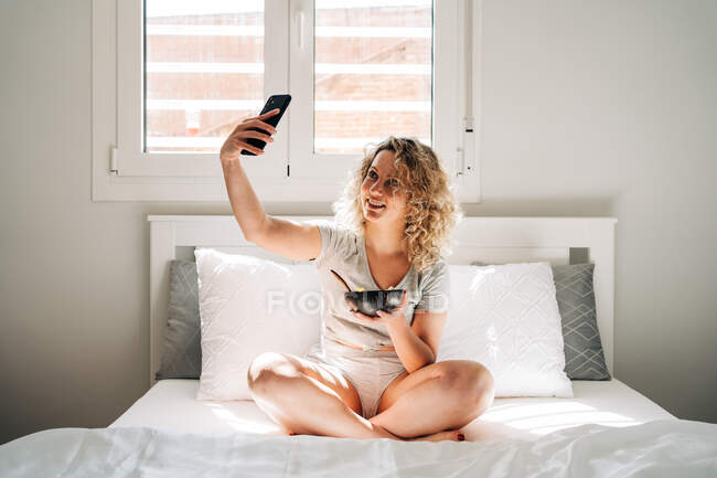 Glad young female in domestic clothes taking selfie pictures of yummy breakfast in bowl while sitting with legs crossed on cozy bed in morning — Photo de stock