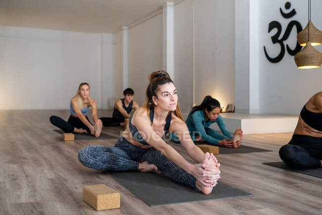 Compañía de personas multiétnicas sentadas en esteras y piernas estiradas en pose Forward Bend durante clase de yoga en estudio - foto de stock