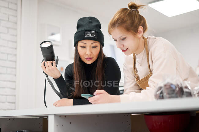 Cheerful female blogger showing cellphone to ethnic girlfriend with professional photo camera while interacting at home — Stock Photo