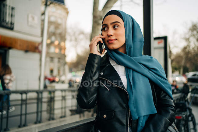 Encantadora mujer musulmana con pañuelo en la cabeza parada en la calle de la ciudad y hablando por teléfono mientras mira hacia otro lado - foto de stock
