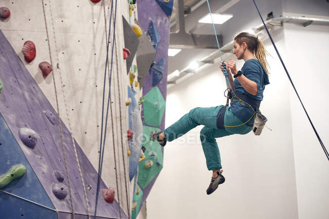 Vista laterale di alpinista donna appesa alle attrezzature di sicurezza durante l'allenamento nel moderno club di boulder — Foto stock